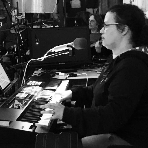A close-up image of a composer working in a studio, surrounded by musical instruments and equipment, conveying the creative process of composing music for a TV series. The focus is on the composer's hands as they interact with the keyboard.
