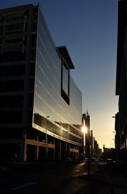A stark photograph of an office building reminiscent of Lumon, but situated in broad daylight amidst a generic corporate park. The image conveys the mundane and anonymous nature of corporate environments contrasting the mysterious interior of Lumon. Focus on clean lines and imposing structure.